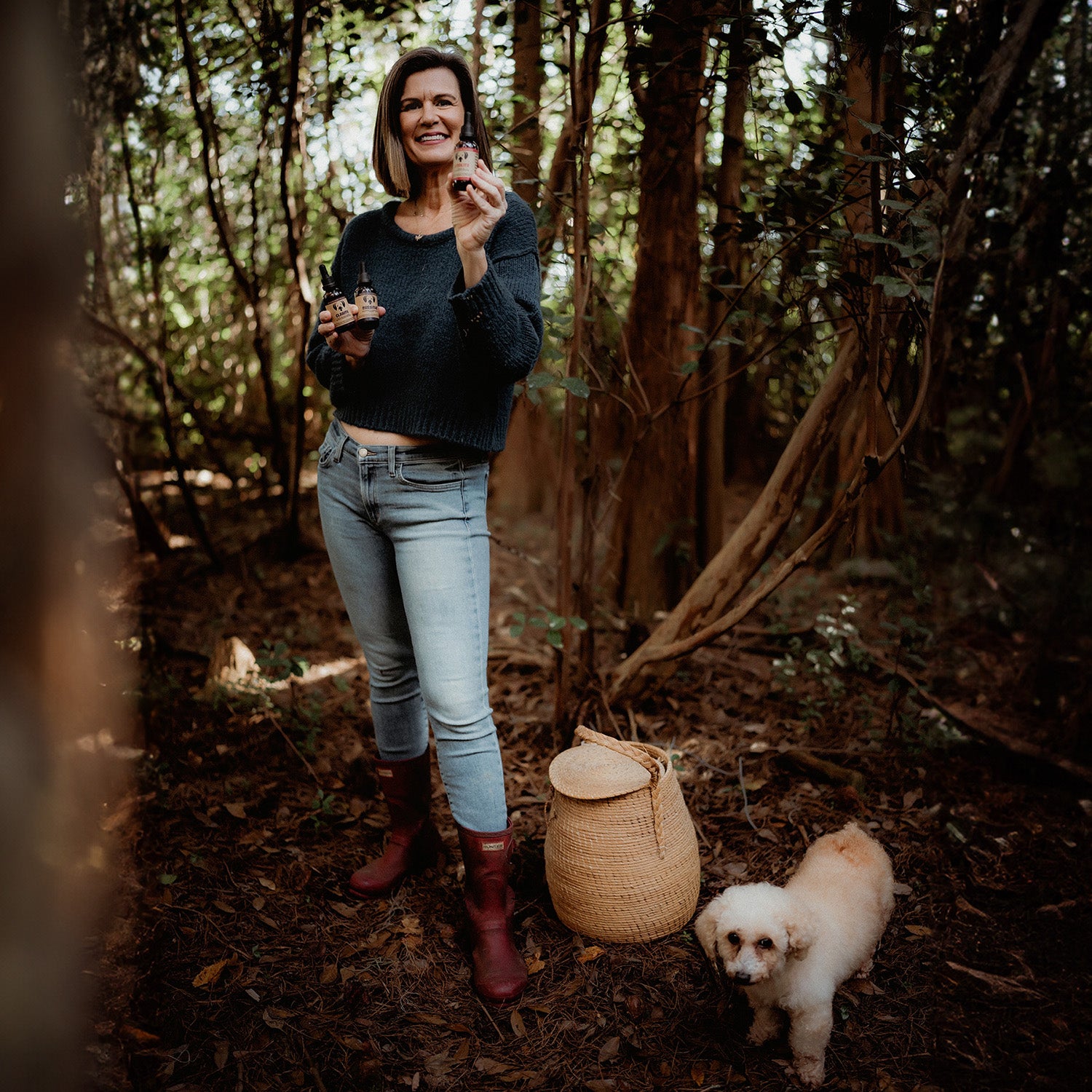 Angela Ardolina founder of MycoDog holding mushroom supplements for dogs in a forest with a small dog and a woven basket on the ground